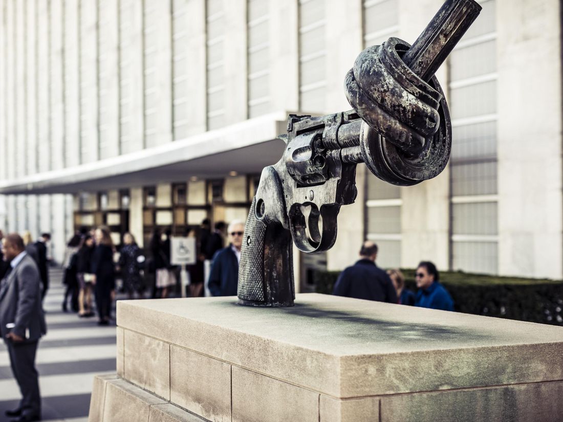 A sculpture of a revolver with a knotted barrel is displayed on a pedestal outside a building, with people gathered nearby.