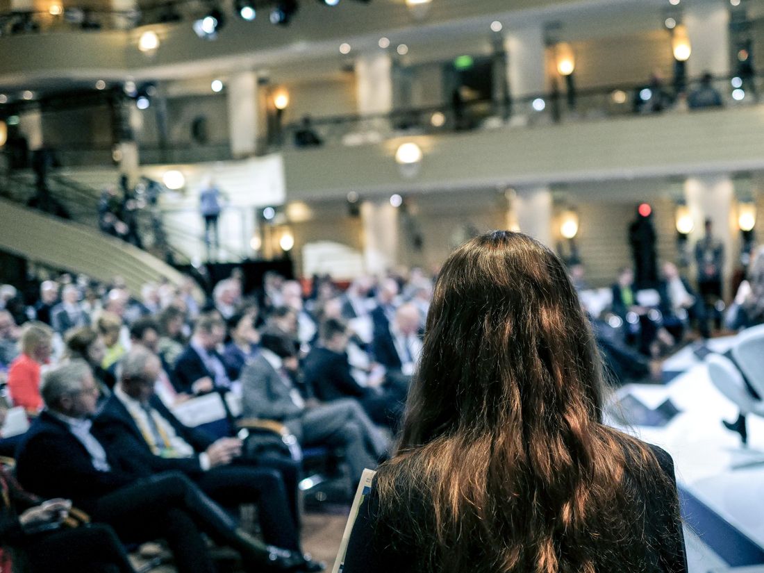 Eine Person mit langen Haaren steht mit dem Rücken zur Kamera, im Hintergrund ist das Publikum der Münchner Sicherheitskonferenz zu sehen.