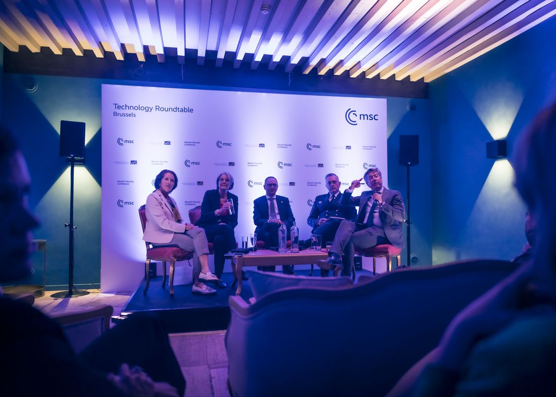 Der Raum ist in ein violettes Licht getaucht. Die Sprecher:innen der Night Cap Session sitzen auf Stühlen, etwas erhöht, auf einer Bühne. Benedikt Franke, Vice-Chair und CEO der Münchner Sicherheitskonferenz ist unter ihnen, hat ein Mikrofon in der einen Hand und deutet mit der anderen Hand mit dem Zeigefinger in das Publikum.