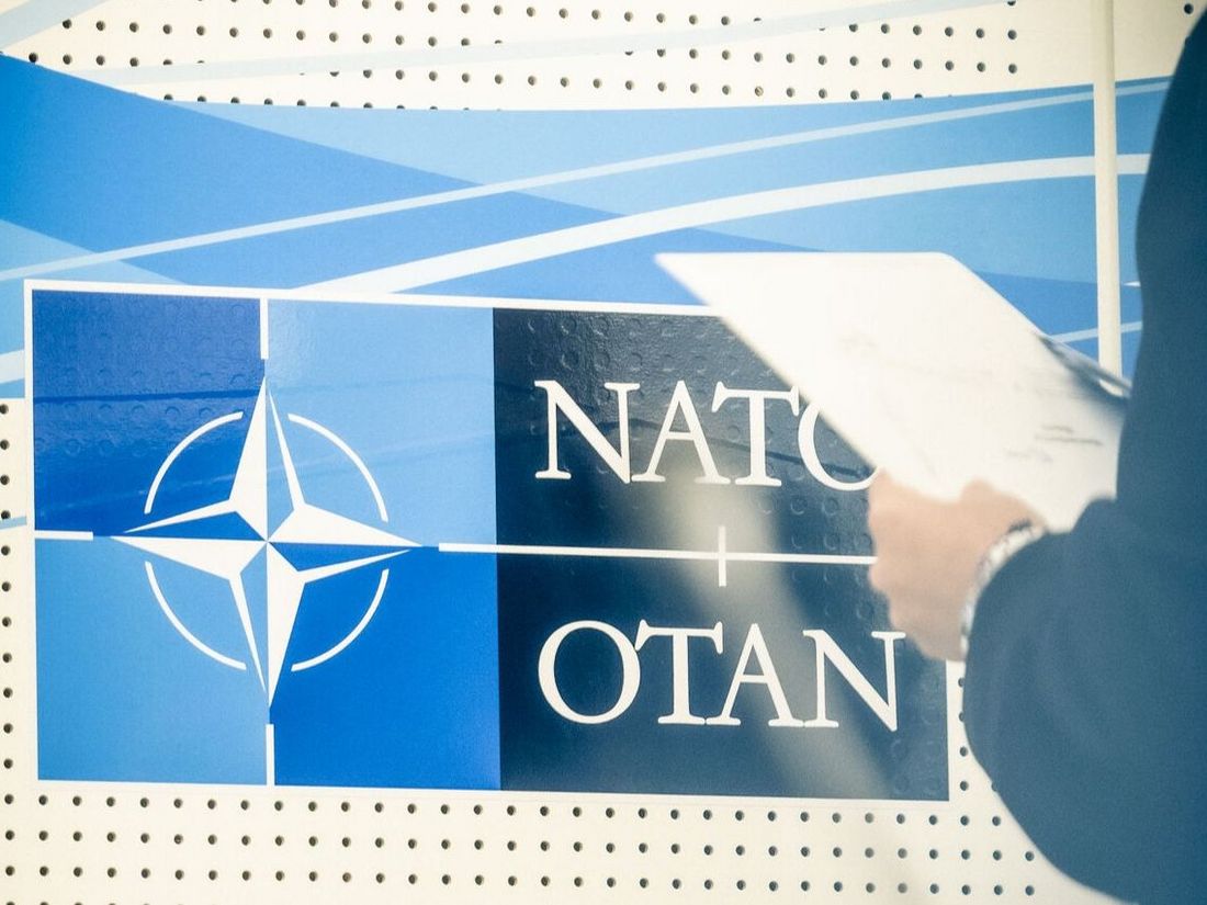 A person with a clipboard stands in front of the NATO logo.