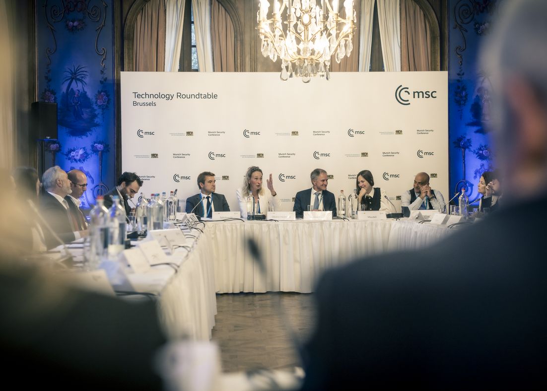 The outlines of two people's shoulders can be seen in the foreground, while the speakers of the second session of the Technology Roundtable can be seen in the background.