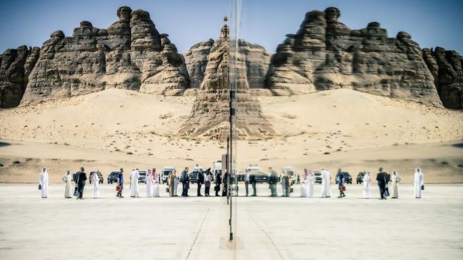 The MLM participants wait in line in front of the location, whose reflective facade mirrors the people and the desert in the background.